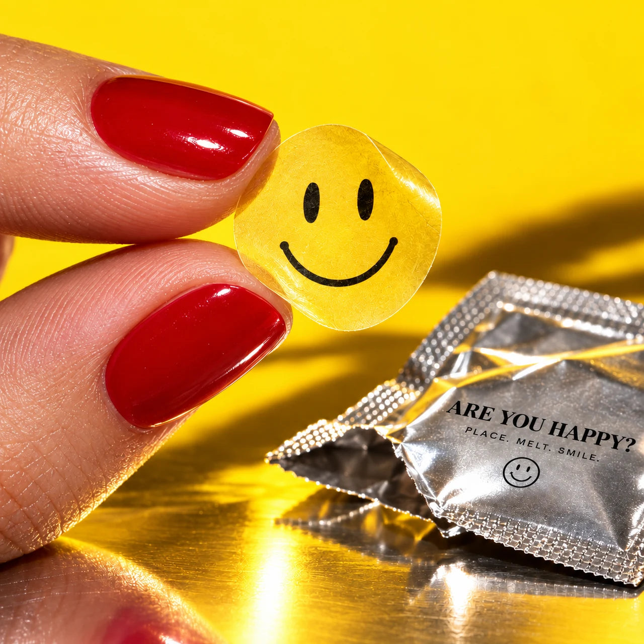 Close-up of a translucent yellow smiley film held between adult fingertips beside an open silver sachet